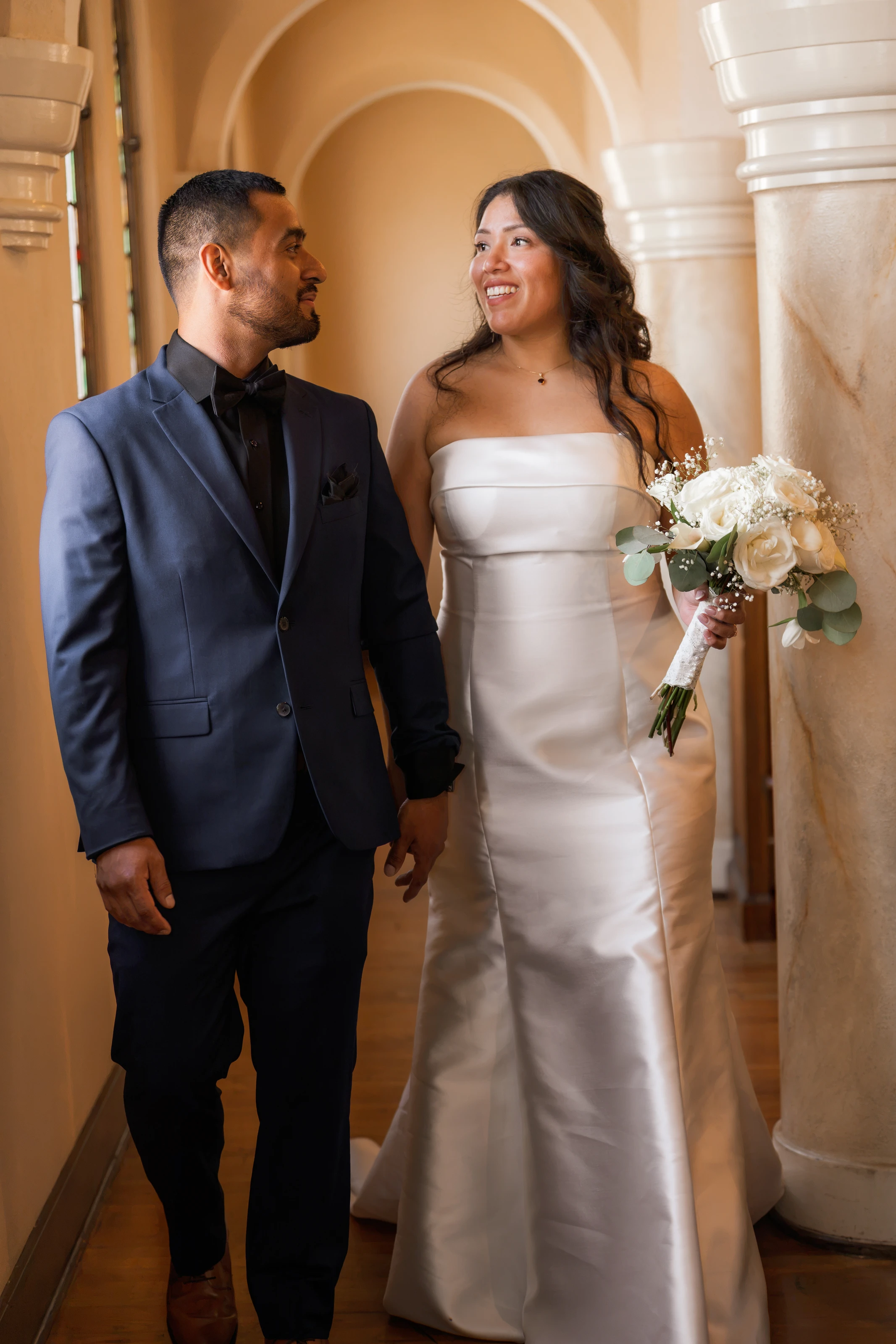 Los novios caminan de la mano por un pasillo con arcos, sonriéndose