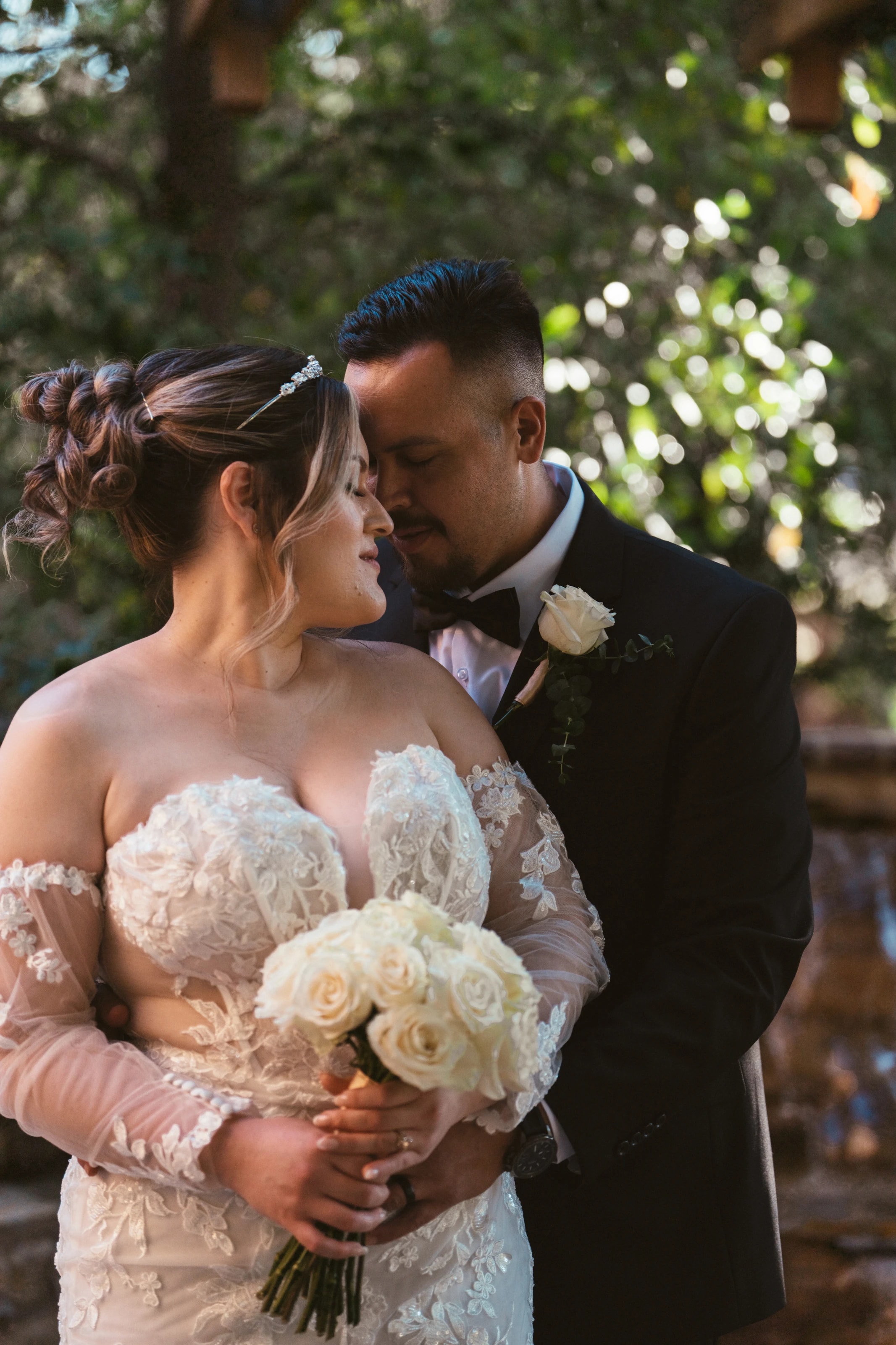 El novio abraza a la novia por detrás, ella con los ojos cerrados y rosas blancas en las manos